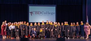 CBD College graduates stand on stage with professors, a screen displaying the college logo behind them.