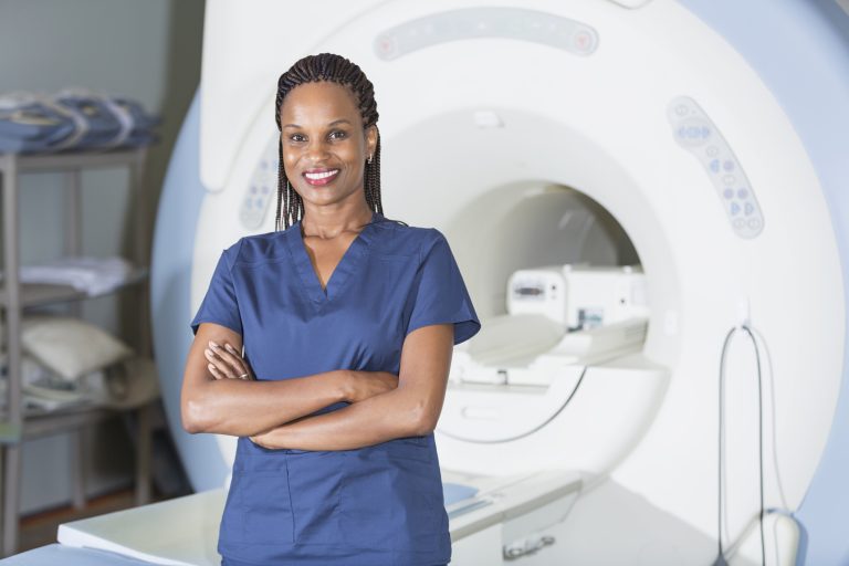 A successful, mature black woman working as a medical professional in a hospital or clinic, standing confidently in front of an MRI scanner, smiling at the camera.