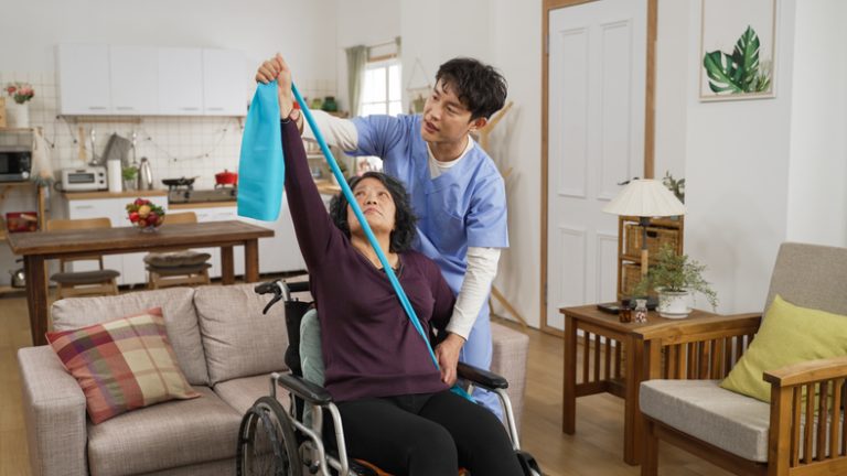 A caring male nurse provides support as a senior female practices using a stretch band for physiotherapy while seated in a wheelchair at home.